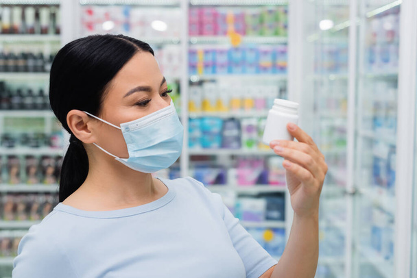 mujer asiática en máscara médica mirando botella con vitaminas en farmacia - Foto, imagen