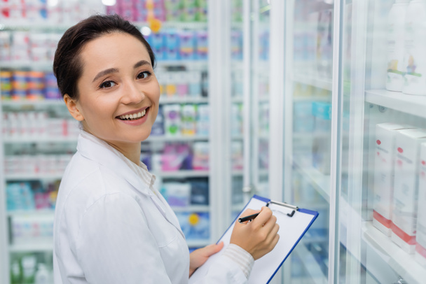 joyful pharmacist looking at camera writing on clipboard  - Foto, Imagem