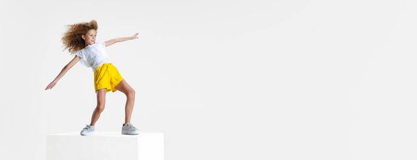 Full-length portrait of smiling girl in casual clothes standing on big box isolated on white studio background. Happy childhood concept. - Фото, изображение