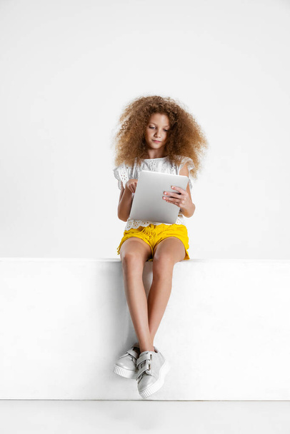 One little cute curly girl in casual clothes sitting on big box with digital tablet, gadget isolated on white studio background. - Fotografie, Obrázek