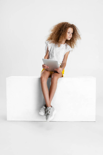 One little cute curly girl in casual clothes sitting on big box with digital tablet, gadget isolated on white studio background. - Photo, Image
