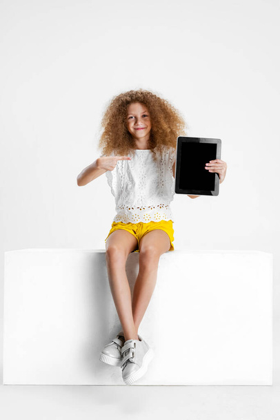 One little cute curly girl in casual clothes sitting on big box with digital tablet, gadget isolated on white studio background. - Foto, Imagem