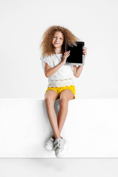 One little cute curly girl in casual clothes sitting on big box with digital tablet, gadget isolated on white studio background. - Foto, Imagem