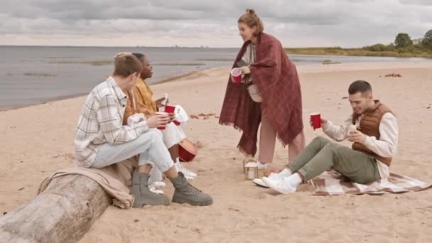 Długie ujęcie pięciu wielonarodowych przyjaciół siedzących na piaszczystej plaży na pikniku z drinkami, a następnie kaukaska kobieta wstaje i zbiera wszystkich za robienie selfie na smartfonie - Materiał filmowy, wideo