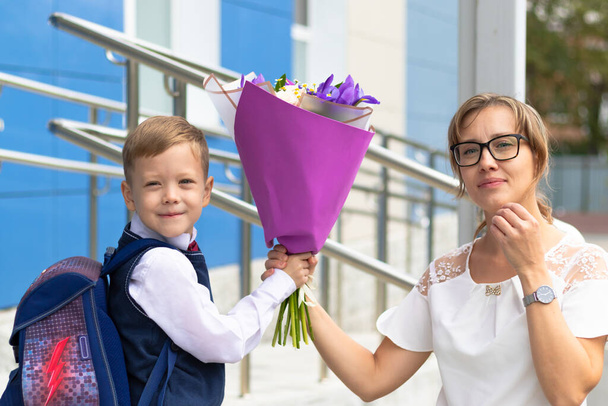 Ένα χαριτωμένο πρωτοκλασάτο αγόρι με σχολική στολή με τη μητέρα του και ένα όμορφο μπουκέτο πολύχρωμα λουλούδια στην αυλή του σχολείου. Εορτασμός την 1η Σεπτεμβρίου. Ημέρα γνώσης. Επιλεκτική εστίαση - Φωτογραφία, εικόνα