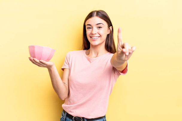 young hispanic woman smiling proudly and confidently making number one. empty bowl concept - Foto, Bild