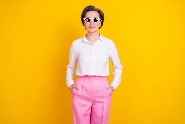 Photo of adorable cool age woman dressed white shirt dark glasses standing arms pockets smiling isolated yellow color background - Zdjęcie, obraz