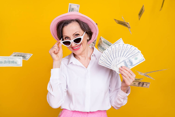 Photo of aged woman happy positive smile hold dollars air fly cash rich isolated over yellow color background - Foto, Imagem