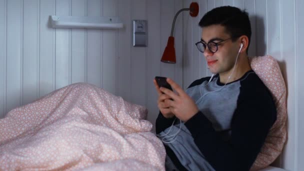Yatakta kulaklıkla uzanan ve geceleri mesajlaşan genç bir adamın yan görüntüsü. - Video, Çekim