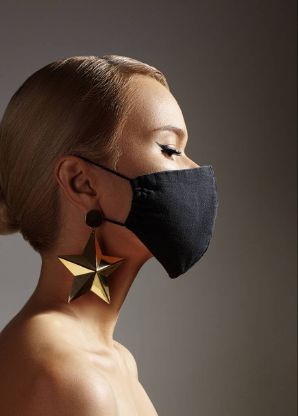 Beautiful Woman with Black Medical Mask on Face. Fashion Eye Liner Make-up. Festive Look with Gold Accessory. Protection Hygiene in Viral Covid-19 Pandemic - Zdjęcie, obraz