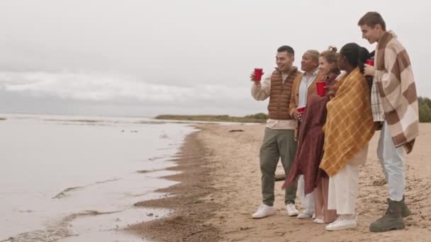 Vista lateral larga de cinco amigos femeninos y masculinos multiétnicos de pie en la costa arenosa, sosteniendo copas de plástico rojo, calentándose con mantas, disfrutando de la vista al mar en el frío día nublado, hablando y sonriendo - Metraje, vídeo