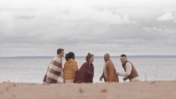 Enfermé de cinq jeunes gens debout sur la côte, étreignant, se réchauffant avec des couvertures, profitant d'une vue sur la mer par temps froid et nuageux, puis se retournant et s'enfuyant, souriant - Séquence, vidéo
