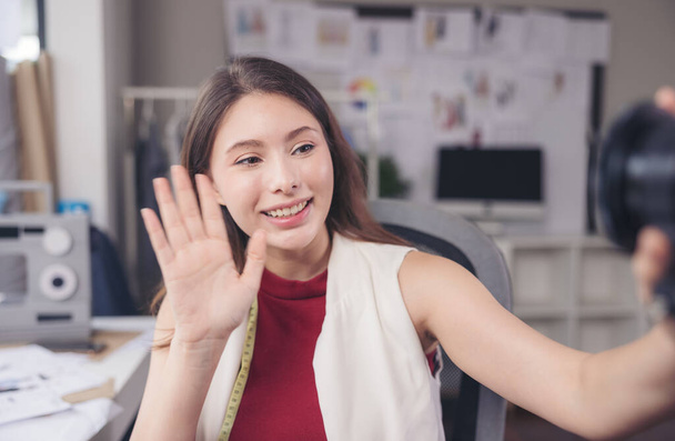 Waving her hand, a fashion designer sits at her workstation in her studio, looks at the camera online present, and communicates through video call. - Foto, immagini