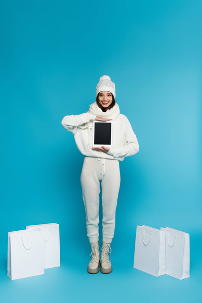 Cheerful woman in warm knitted clothes holding digital tablet near shopping bags on blue background  - Foto, afbeelding