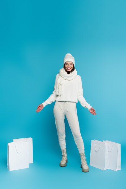 Positive woman in knitted clothes pointing at shopping bags on blue background  - Фото, изображение