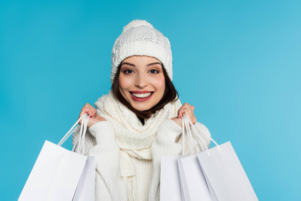 Happy young woman in white hat and scarf holding shopping bags isolated on blue  - Fotoğraf, Görsel