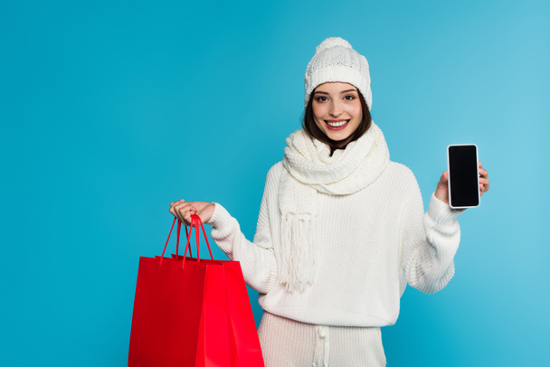 Young woman in warm clothes holding smartphone with blank screen and shopping bags isolated on blue  - Foto, afbeelding