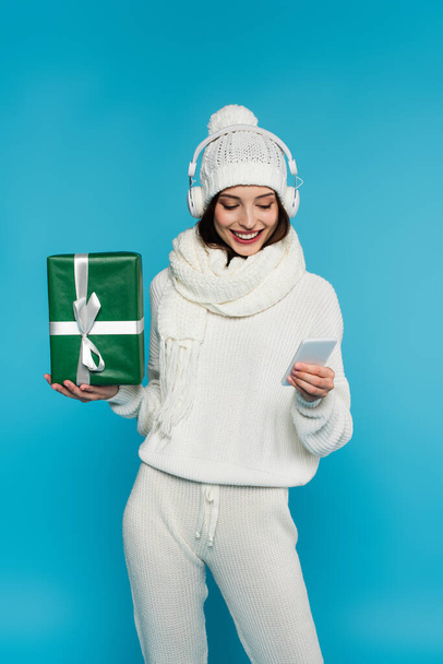 Smiling woman in headphones and warm clothes using cellphone and holding gift isolated on blue  - Foto, Imagem