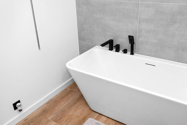 A modern, free standing wall mounted bathtub with a black matt tap, standing in a bathroom lined with ceramic tiles. - Valokuva, kuva