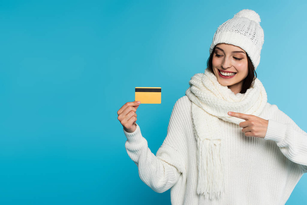Mujer positiva en suéter cálido y sombrero apuntando a la tarjeta de crédito aislado en azul  - Foto, Imagen
