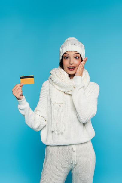 Excited woman in knitted clothes holding hand near cheek and credit card isolated on blue  - Foto, immagini