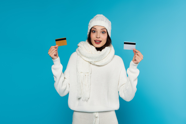 Astonished woman in knitted clothes holding credit cards isolated on blue  - Foto, afbeelding