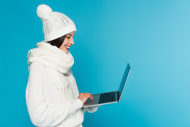 Side view of smiling freelancer in knitted hat and scarf using laptop isolated on blue  - Valokuva, kuva