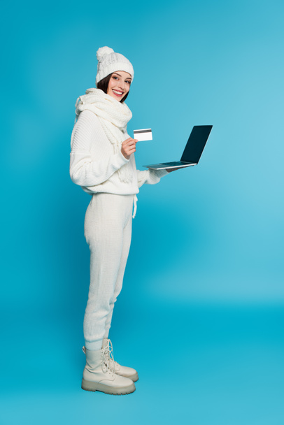 Full length of smiling woman in knitted clothes holding credit card and laptop with blank screen on blue background  - 写真・画像