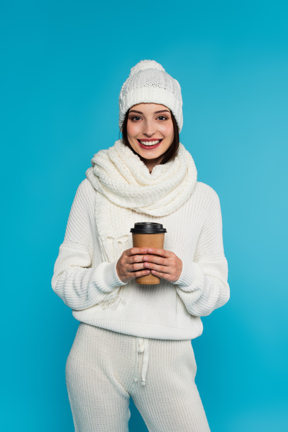 Pretty woman in knitted scarf and hat holding coffee to go isolated on blue  - Фото, изображение