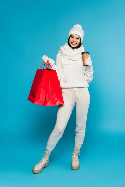 Mulher positiva em camisola de malha e luvas segurando café para ir e sacos de compras no fundo azul  - Foto, Imagem