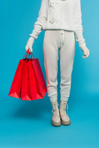 Cropped view of woman in knitted clothes and gloves holding red shopping bags on blue background  - Foto, Bild