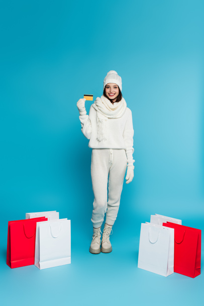 Smiling woman in warm clothes and gloves holding credit card near shopping bags on blue background  - Foto, Imagen