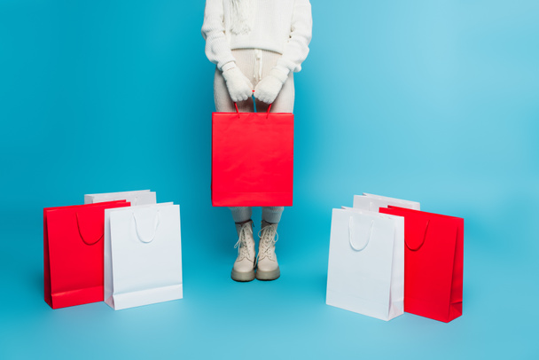 Cropped view of woman in sweater and gloves holding shopping bag near purchases on blue background  - Foto, Bild
