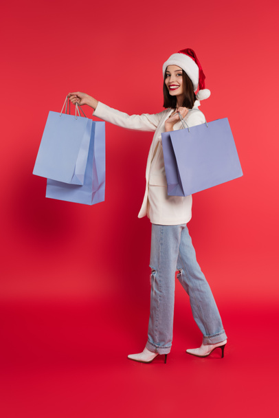 Cheerful woman in jacket and santa hat holding shopping bags on red background  - Foto, immagini