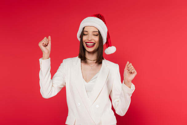 Excited woman in santa hat and white jacket showing yes gesture isolated on red  - 写真・画像