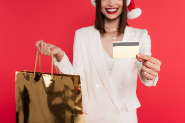 Cropped view of blurred woman in santa hat holding shopping bag and credit card isolated on red  - Photo, image