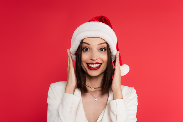 Excited woman in santa hat and white jacket looking at camera isolated on red  - 写真・画像