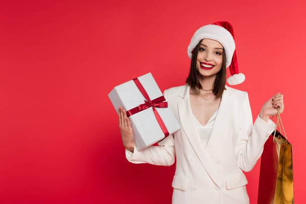 Happy woman in santa hat and white jacket holding shopping bag and gift box isolated on red  - Foto, Imagem