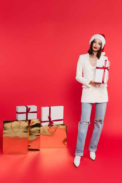 Pretty woman in santa hat holding gift near shopping bags on red background  - Photo, Image