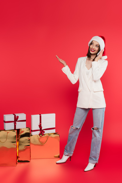 Smiling woman in santa hat talking on smartphone near gifts and shopping bags on red background  - Foto, Bild
