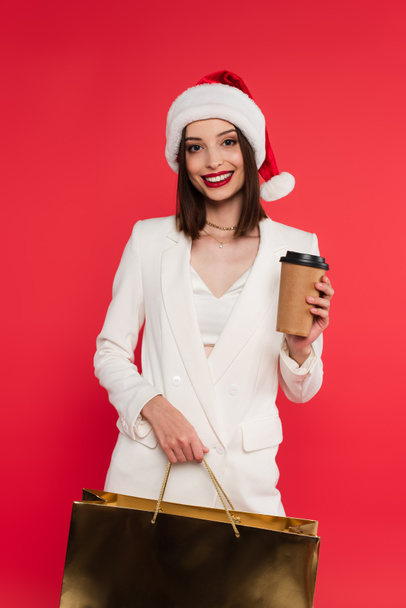 Stylish woman in santa hat holding shopping bag and coffee to go isolated on red  - Photo, Image