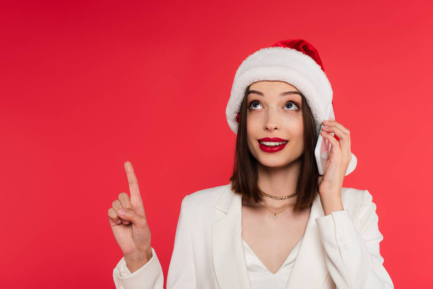 Smiling woman with red lips and santa hat talking on smartphone and pointing with finger isolated on red  - Фото, изображение