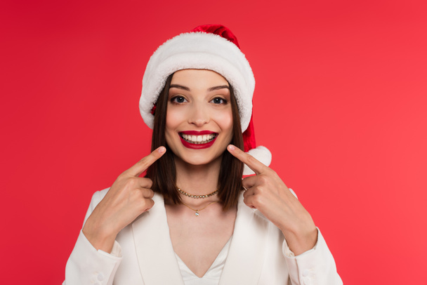Smiling woman in santa hat pointing at teeth isolated on red  - Foto, Imagem