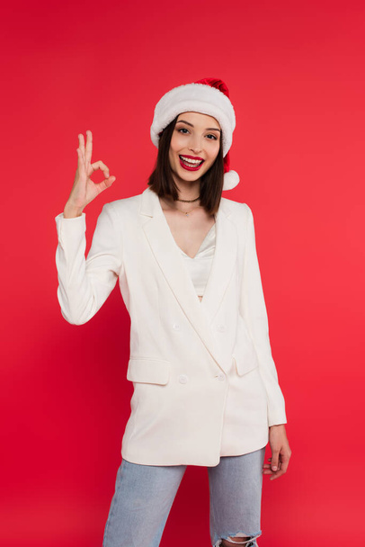 Stylish woman in santa hat showing okay gesture isolated on red  - Foto, Bild