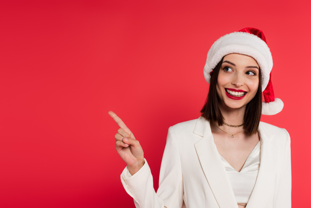 Cheerful woman in santa hat pointing with finger isolated on red  - Φωτογραφία, εικόνα
