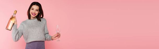 Positive woman holding bottle of champagne and glass isolated on pink, banner  - 写真・画像
