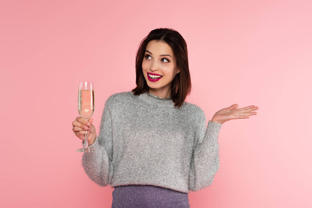 Улыбающаяся женщина в свитере держит бокал с шампанским и указывает рукой, изолированной на розовый  - Фото, изображение