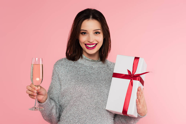 mooie vrouw met champagne en geschenk op zoek naar camera geïsoleerd op roze  - Foto, afbeelding