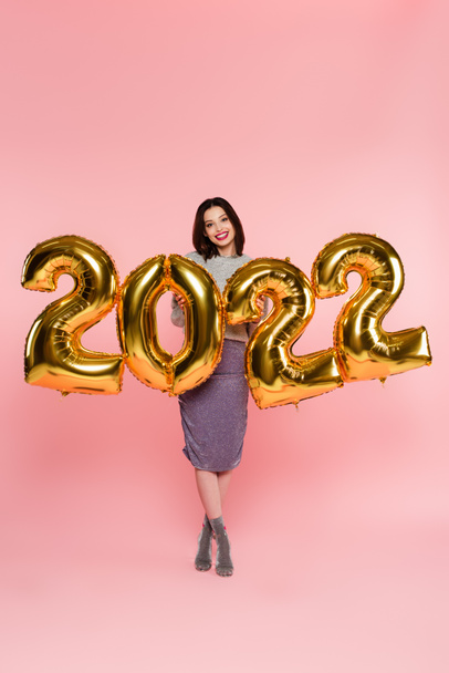 ピンクの背景に2022年の数字の形で風船の近くに笑顔スタイリッシュな女性 - 写真・画像