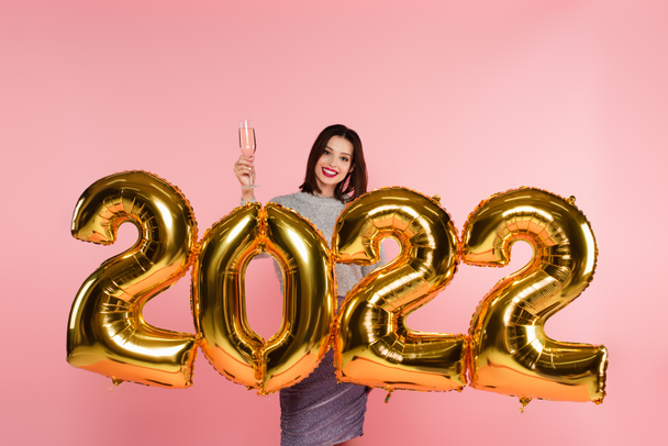 Smiling woman holding glass of champagne near balloons in shape of 2022 isolated on pink  - Photo, Image
